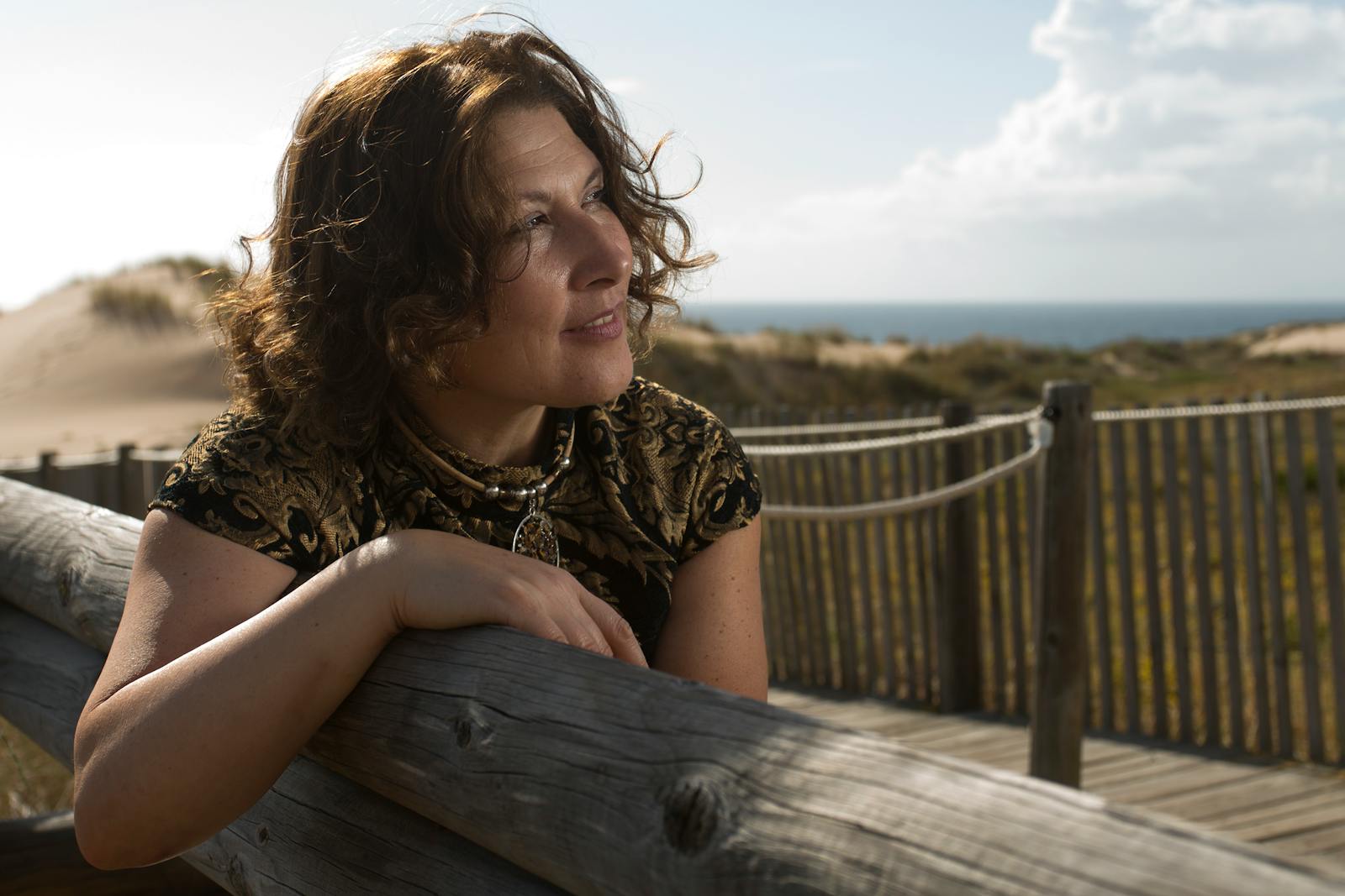 A woman leans on a wooden fence, gazing at the ocean on a sunny day.